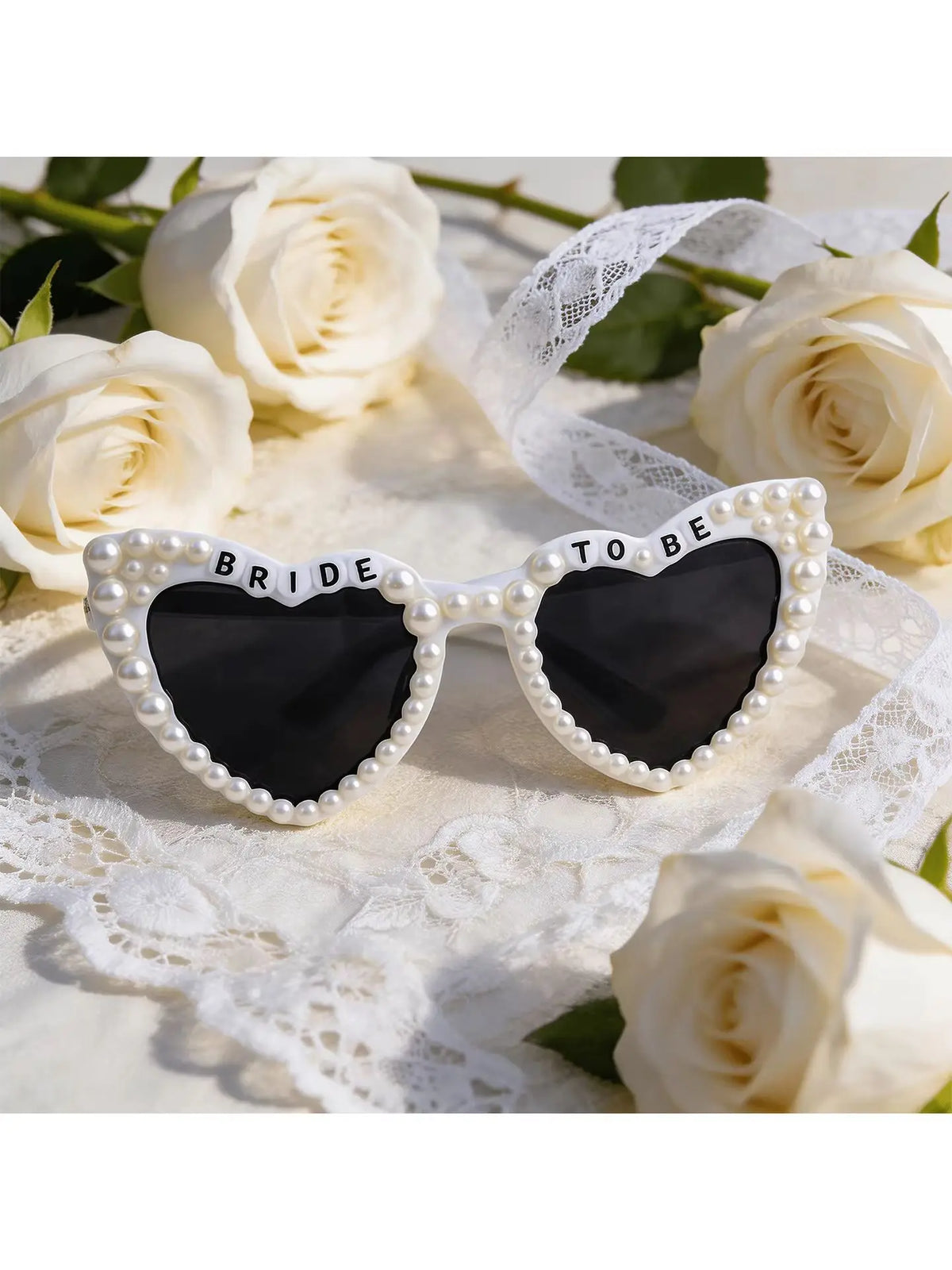 A pair of white pearl-rimmed sunglasses with a heart design and the words bride to be set on a white lace fabric surrounded b