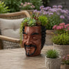A terracotta artistic face planter pot with a carved facial expression sits on a wooden table.