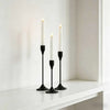 A set of three white taper candles in black holders displayed on a white shelf.