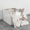 A stack of white faux marble self-adhesive wall tiles with brown veining displayed on a gray floor.