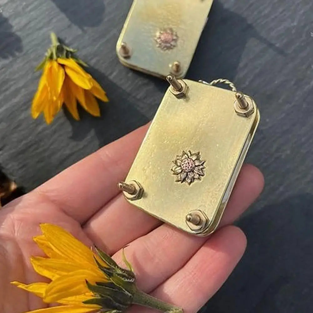 A hand holds a small gold-toned metal flower press kit with a dried yellow flower attached.