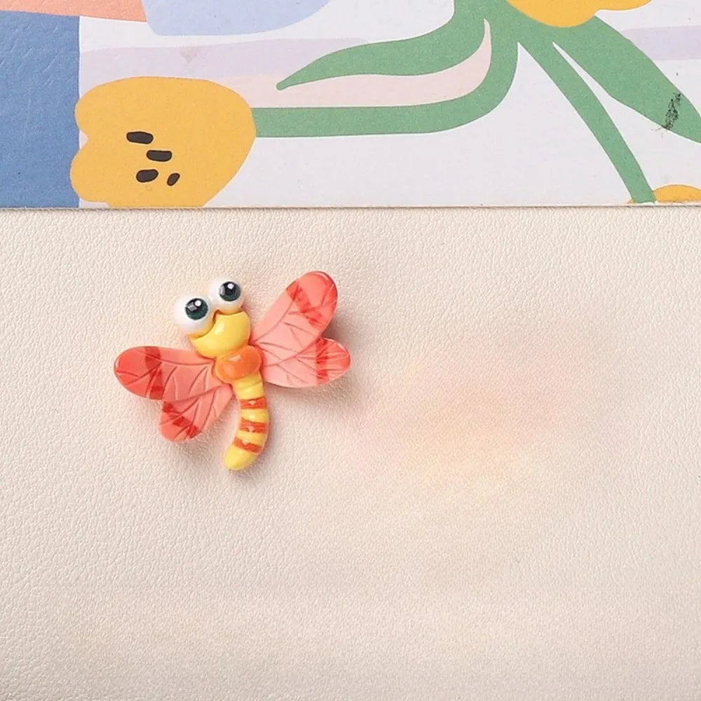 A small, orange and pink resin insect magnet shaped like a butterfly resting on a light beige surface.