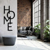 A black metal word art sign spelling HOME mounted on a white wall next to a gray ottoman.