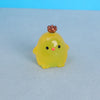 A small, yellow resin chicken ornament with a brown comb and black eyes resting on a gray surface.