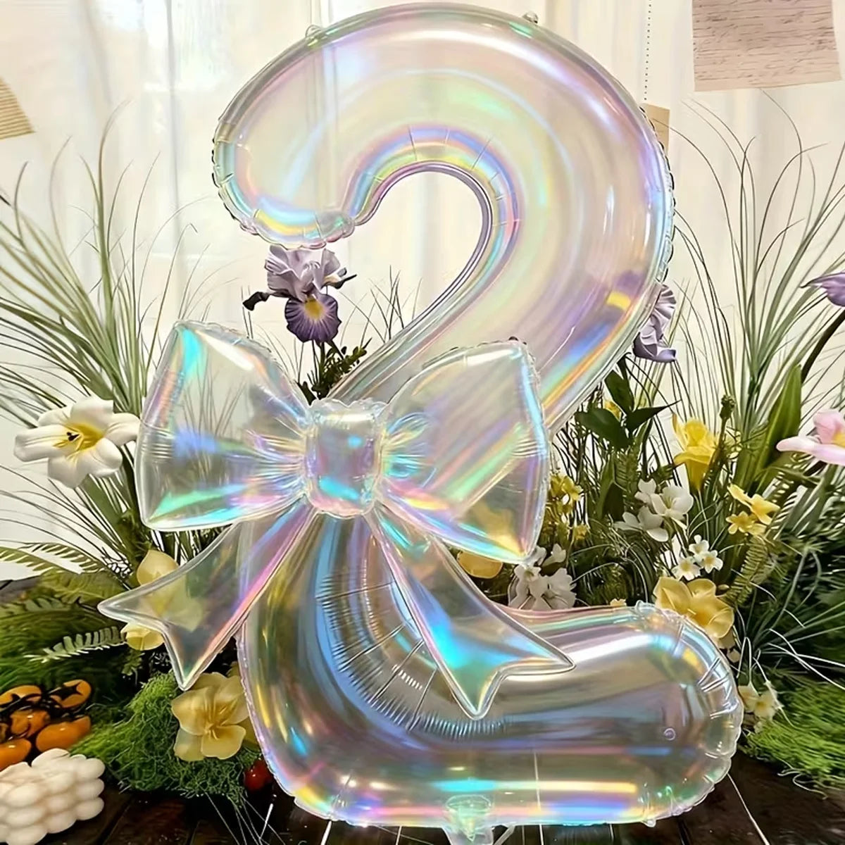 A large, iridescent, transparent number 2 balloon tied in a bow, surrounded by green foliage and white flowers.