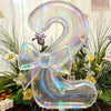 A large, iridescent, transparent number 2 balloon tied in a bow surrounded by green foliage and white flowers.