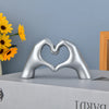 Silver heart shape sculpture made of hand gestures resting on a gray box with yellow flowers in the background