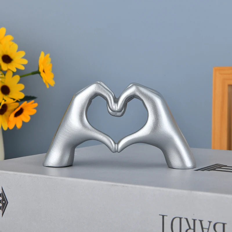 Silver heart shape sculpture made of hand gestures resting on a gray box with yellow flowers in the background