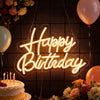 A glowing neon sign spelling Happy Birthday is displayed against a dark background with flowers and balloons.