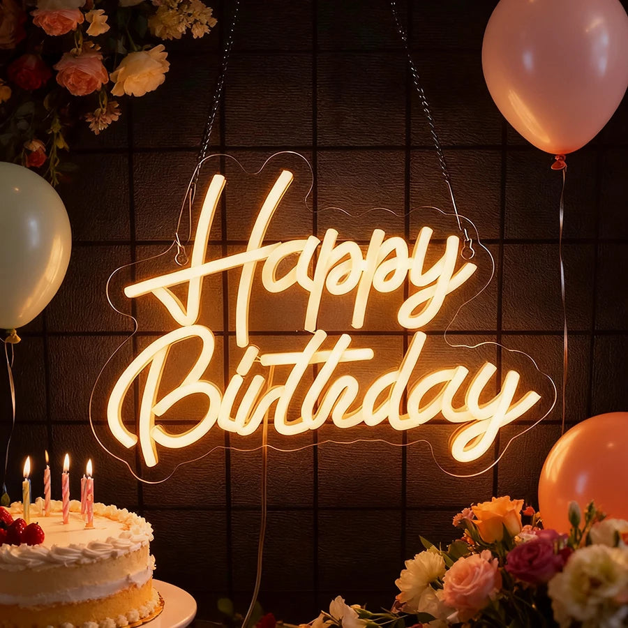 A glowing neon sign spelling Happy Birthday is displayed against a dark background with flowers and balloons.