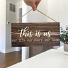 A rustic wooden wall sign with white script text reading This is us our life our story our home, held by a hand.
