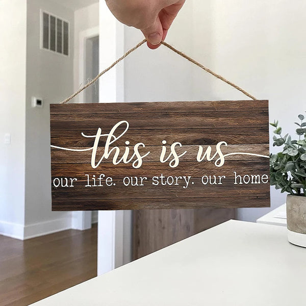 A rustic wooden wall sign with white script text reading This is us our life our story our home, held by a hand.