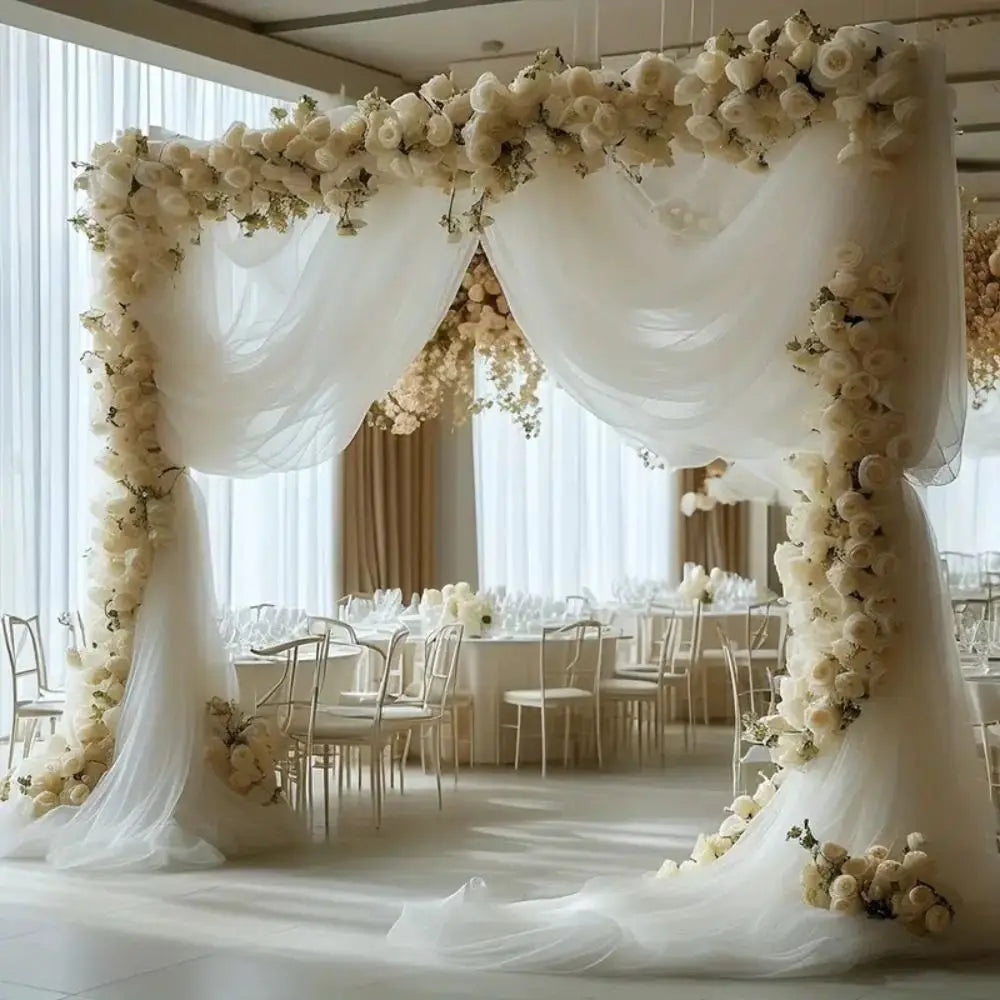 A white sheer tulle fabric backdrop decorated with white roses and draped fabric for a wedding or event setting.