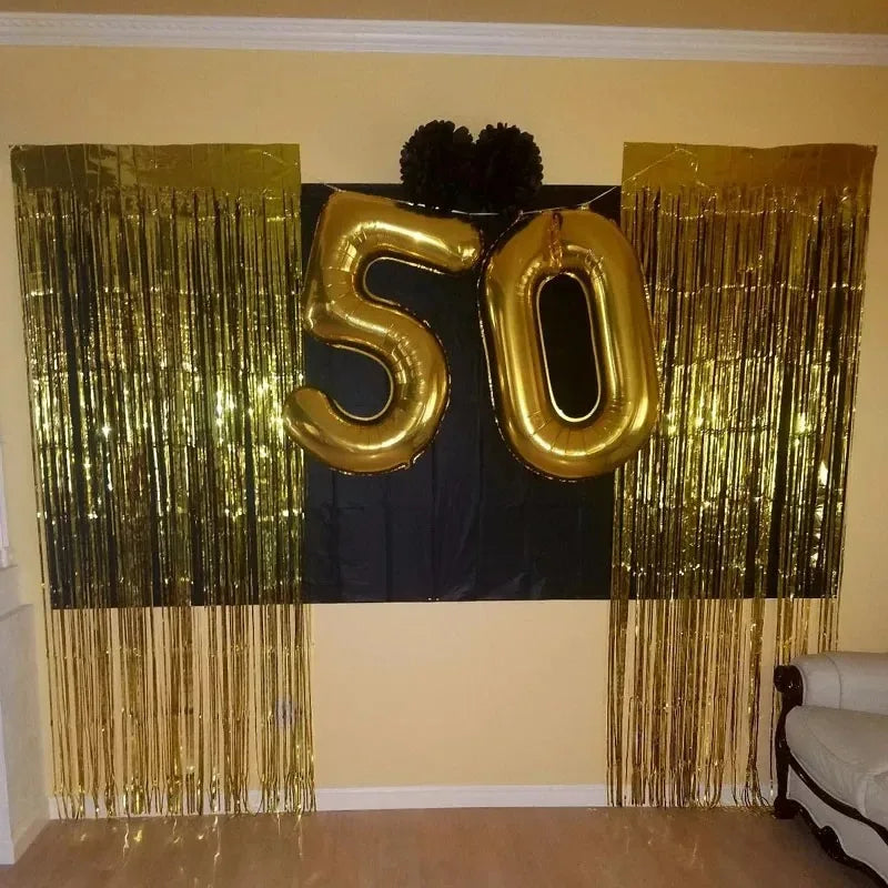 A decorative display featuring large gold foil number balloons '50' flanked by shimmering gold fringe curtains on a wall.