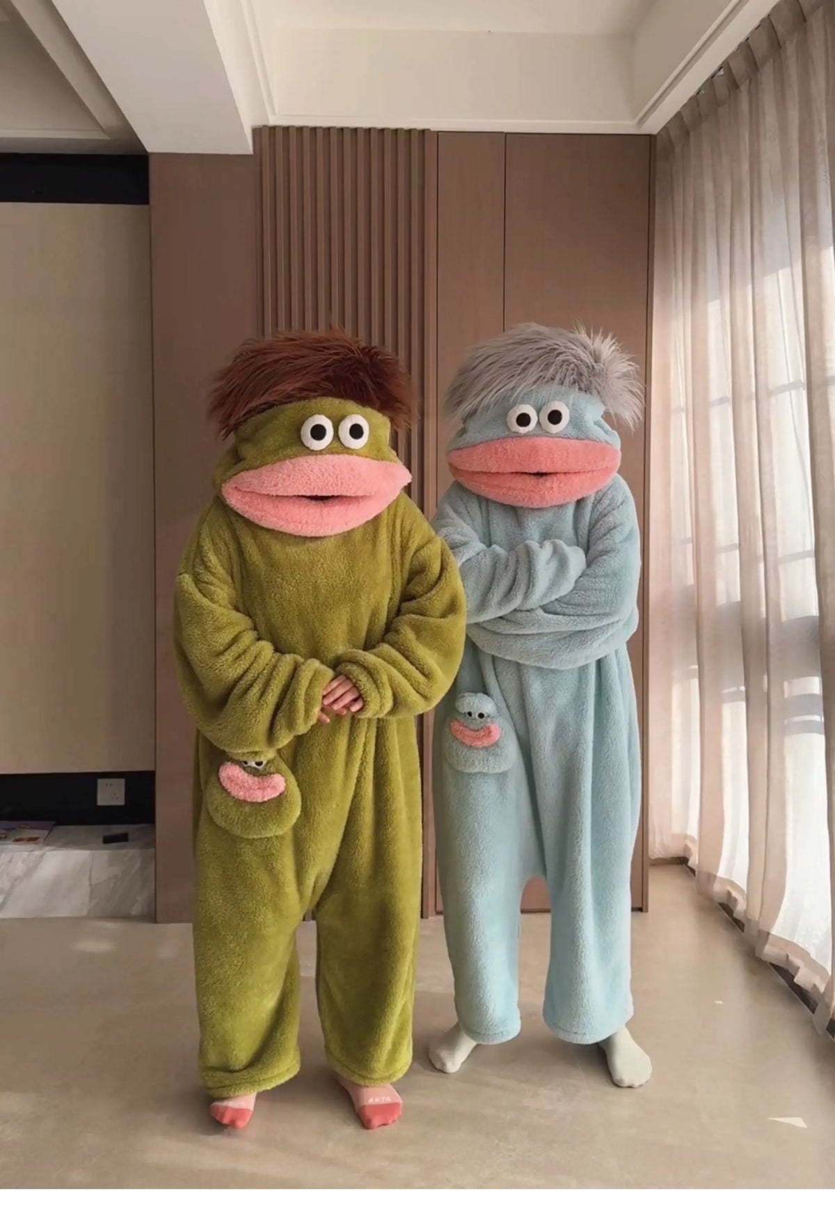A pair of fuzzy green and blue monster onesie pajamas for couples standing in a room.