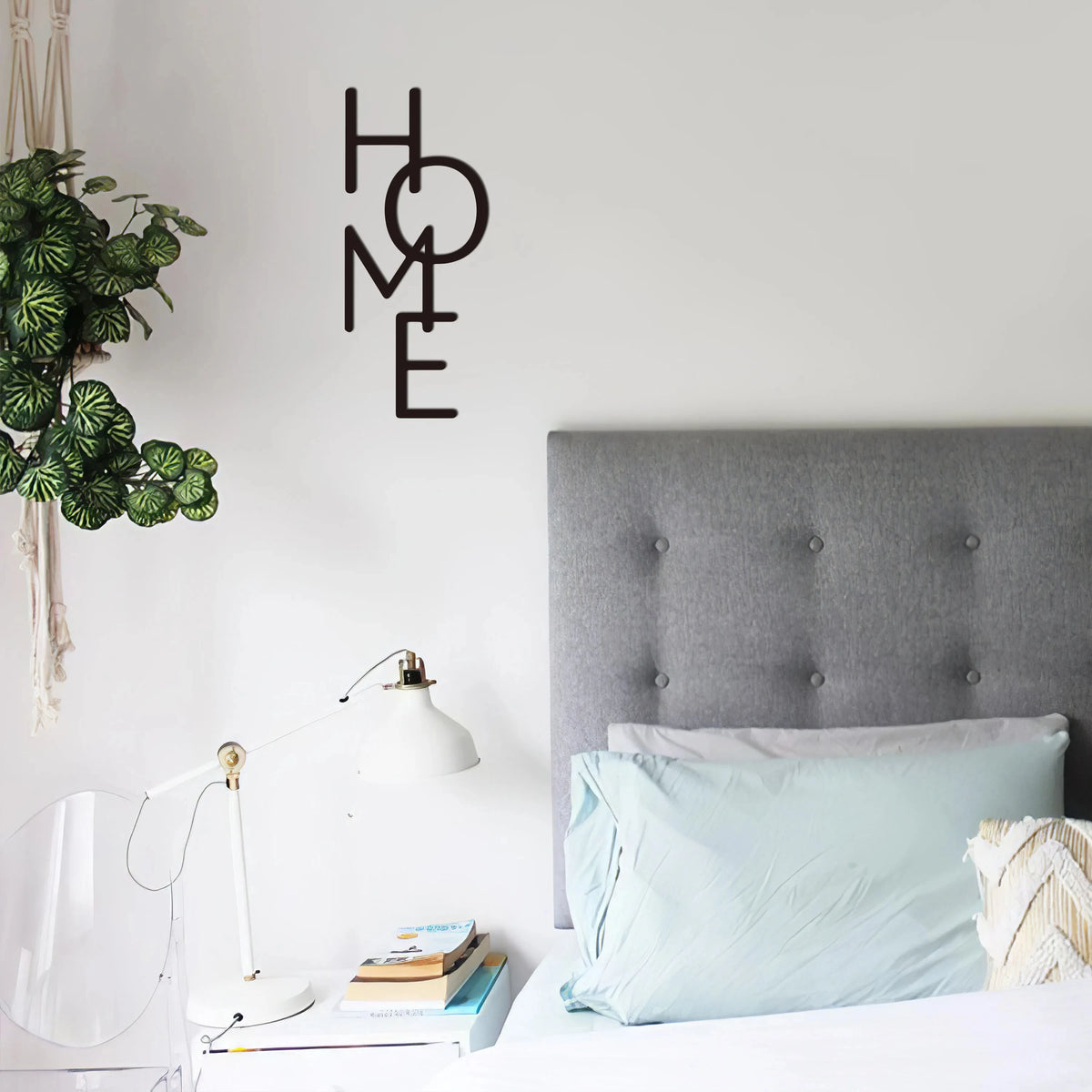 A bedroom scene featuring a metal word art sign spelling HOME above a bed with light blue pillows and a modern lamp.