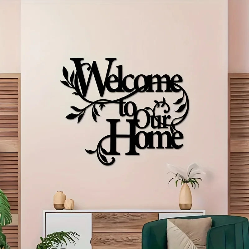A black metal sign with the text Welcome to Our Home mounted on a light pink wall.