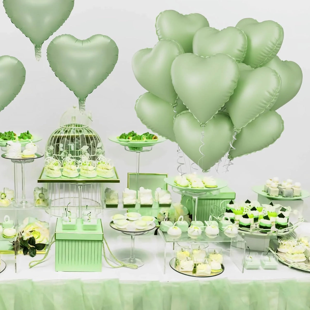 A display of light green heart-shaped foil balloons and various green desserts on a white table.