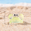 A small, translucent yellow resin crab decor with large eyes resting on a sandy beach.