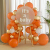 A large balloon arch made of orange and gold latex balloons shaped like a baby, displayed on a wooden stand.