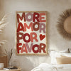 A framed canvas print featuring the text MORE AMOR FOR FAVOR in bold, layered red and pink letters on a light background.
