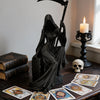 A black gothic female grim reaper figurine holding a scythe, displayed on a dark wooden surface with tarot cards.