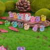 A collection of luminous mini duck figurines in various pastel colors resting on mossy green grass with pinecones and leaves.