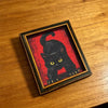 A framed picture of a black cat with yellow eyes on a red background, displayed on a wooden surface.