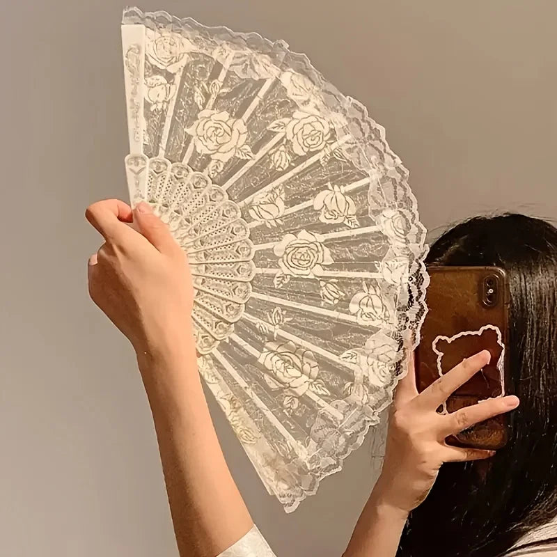 A hand holds an elegant lace rose folding fan with intricate floral patterns against a neutral background.