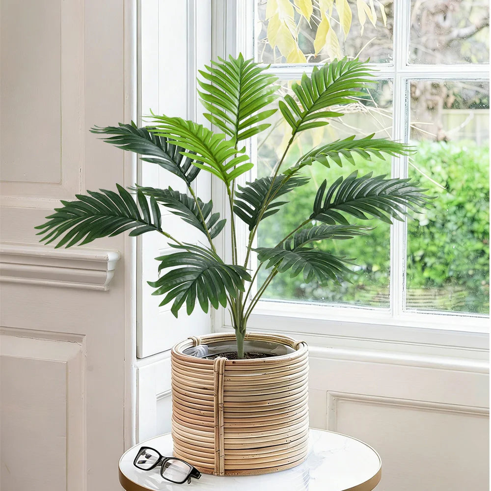 An artificial palm tree in a woven basket sits on a white table next to a window.