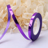 A roll of iridescent purple satin balloon ribbon lying on a white surface next to white flowers.