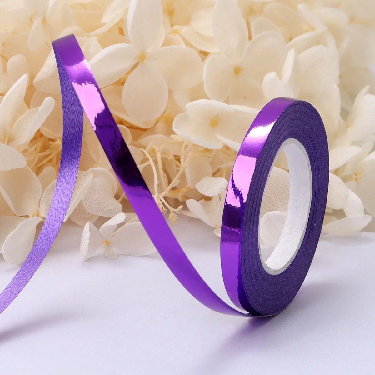A roll of iridescent purple satin balloon ribbon lying on a white surface next to white flowers.