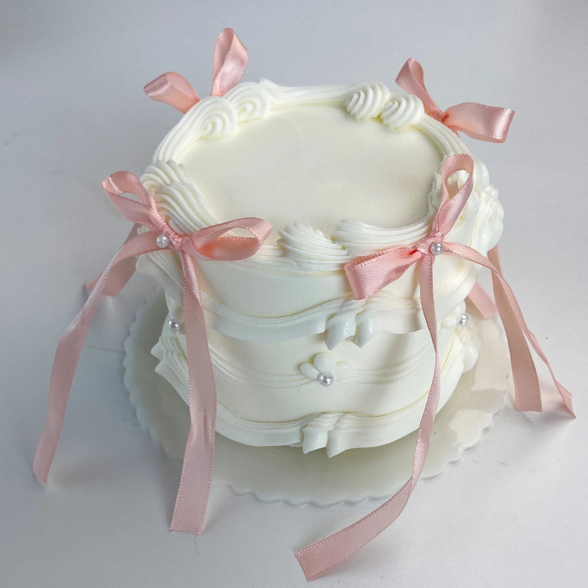 A white decorative fake cake with pink ribbons and pearl accents on a white pedestal platter.