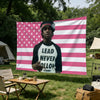A person wearing a black shirt with white text stands in front of a pink and white striped flag banner.