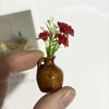 A hand holds a small, brown ceramic vase with artificial red flowers against a white background.