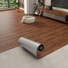 A roll of 3D textured wood grain self-adhesive flooring film placed on a light gray floor next to wooden furniture.