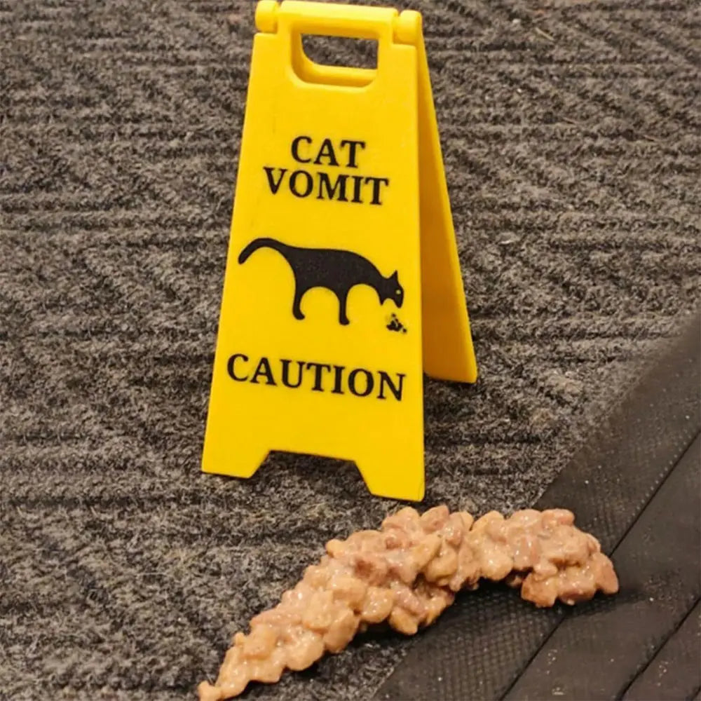 A yellow caution sign for cat vomit placed next to a clump of brownish, semi-solid material on a dark surface.