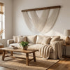 A bohemian macrame wall tapestry hangs above a beige sofa and wooden coffee table in a bright living room.