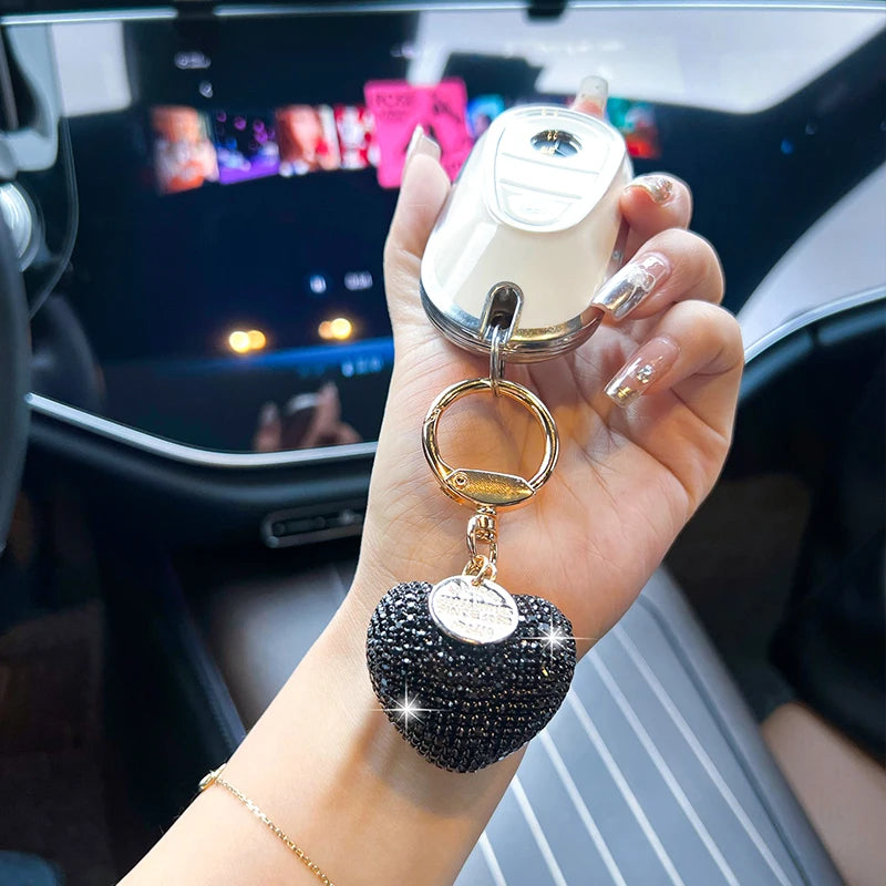 A hand holds a gold-toned keychain featuring a peach heart crystal charm and a silver locket.