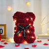 A dense red artificial rose teddy bear illuminated by warm string lights, resting on a white surface with scattered red petal