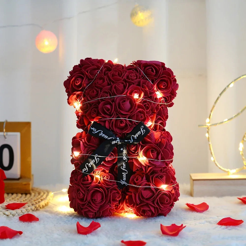 A dense red artificial rose teddy bear illuminated by warm string lights, resting on a white surface with scattered red petal