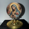 A round religious medal featuring the Virgin Mary with hands clasped in prayer, set on a golden base.