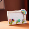 A white photo frame decorated with pink and blue fabric flowers and textured blocks, displayed on a wooden surface.
