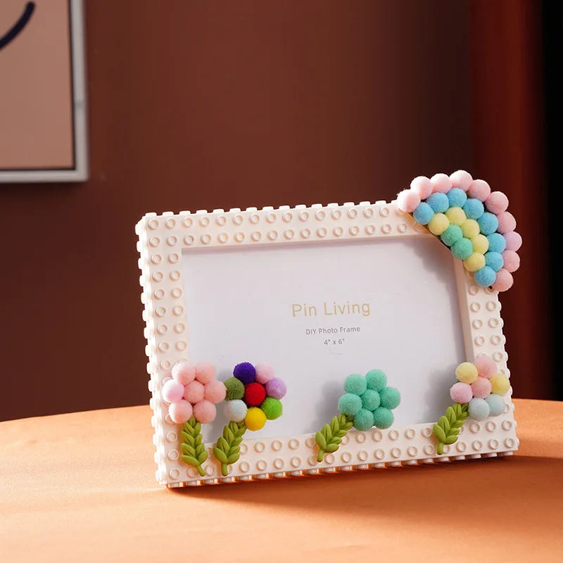 A white photo frame decorated with pink and blue fabric flowers and textured blocks, displayed on a wooden surface.