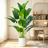 A large artificial Bird of Paradise plant in a white pot sits on a wooden table next to a chair.