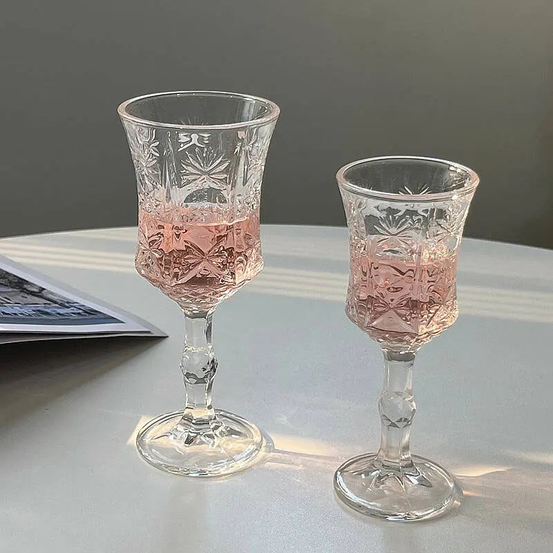 A set of two vintage French-style high-footed wine glasses filled with pink liquid on a white surface.