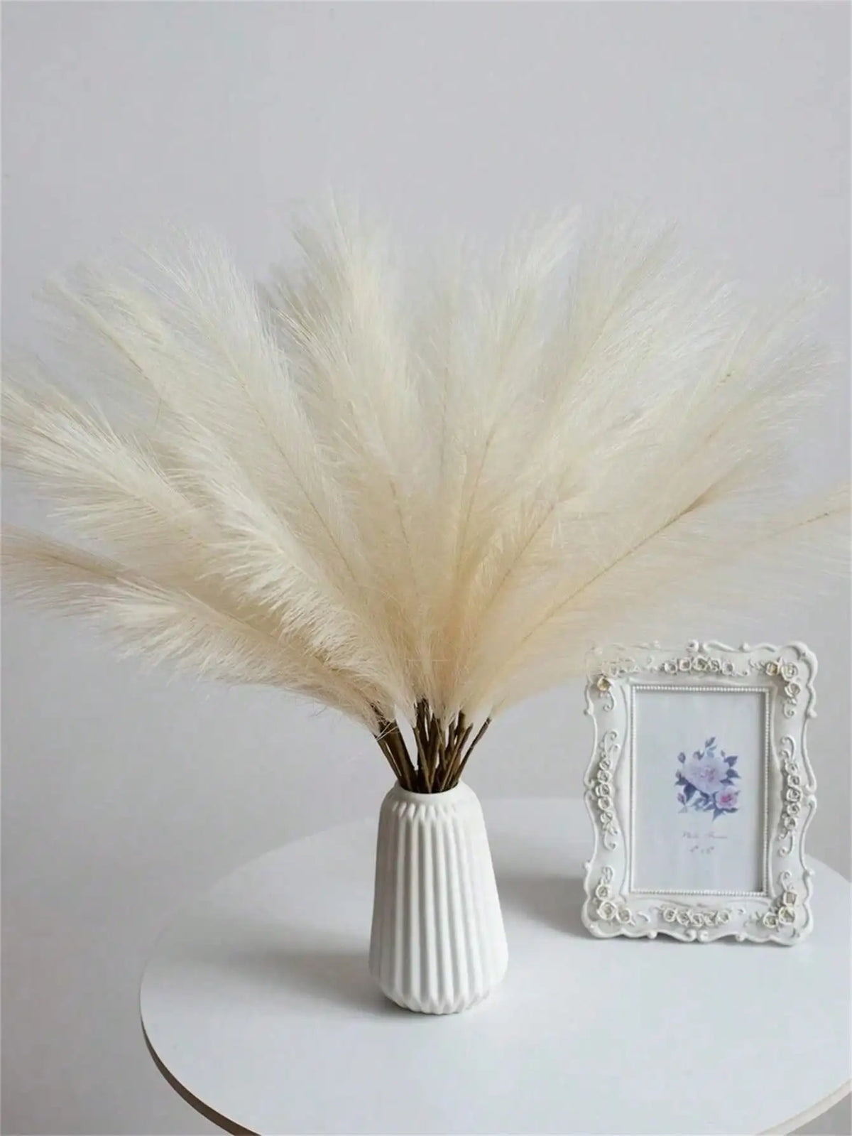 A bundle of fluffy, cream-colored pampas grass stems in a white vase next to a framed floral print.