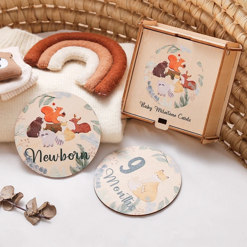 A set of wooden baby milestone cards featuring a newborn theme, displayed next to a wooden keepsake box and a cinnamon-colore