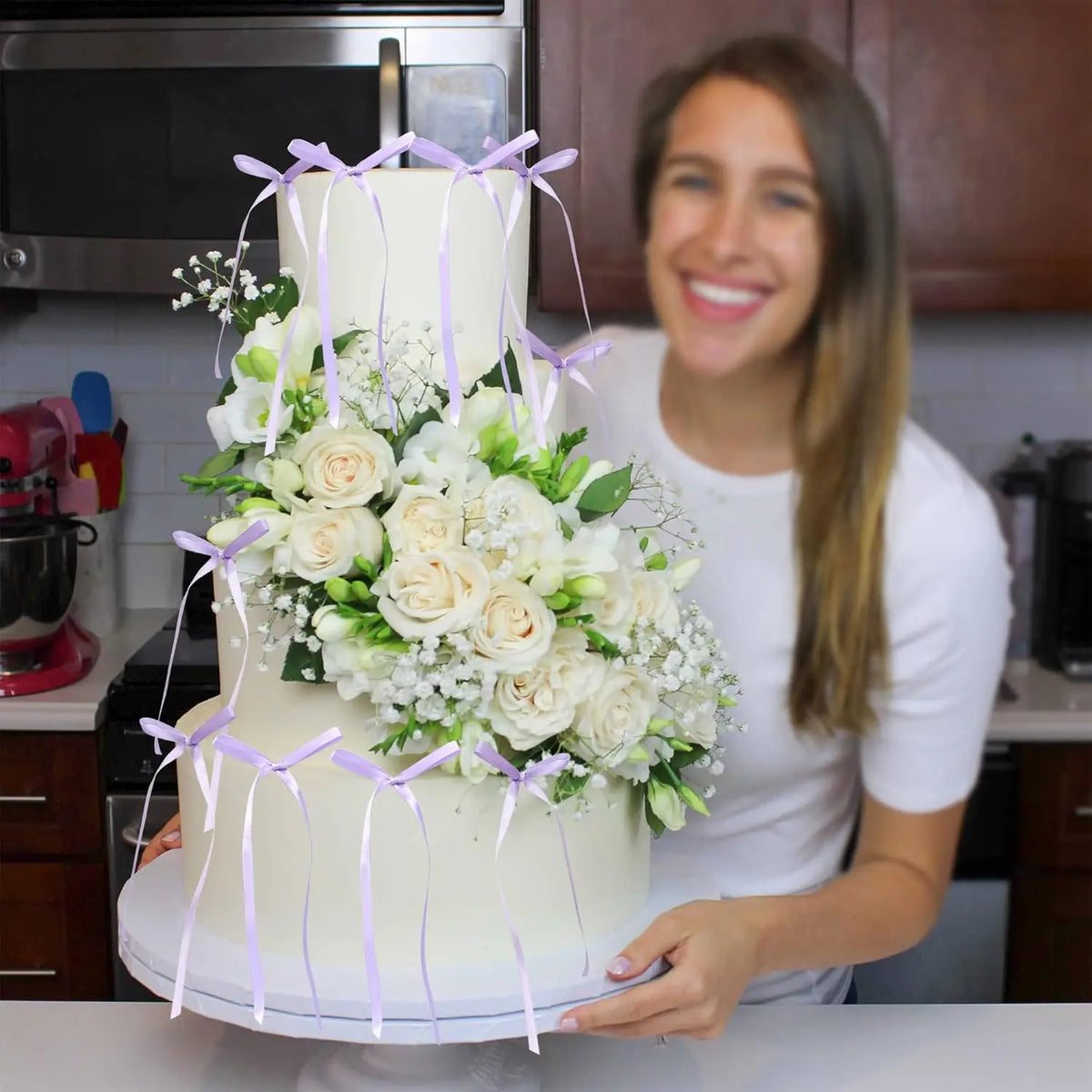 A woman holds an elegant white cake decorated with white roses and purple ribbon bows in a kitchen setting.