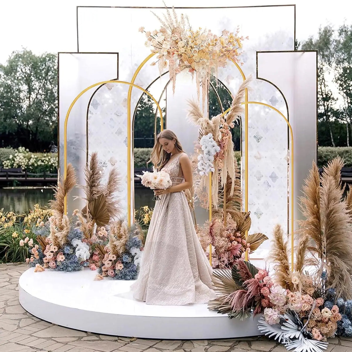 A wedding backdrop featuring a gold metal arched frame decorated with dried floral arrangements and a woman in a flowing gown